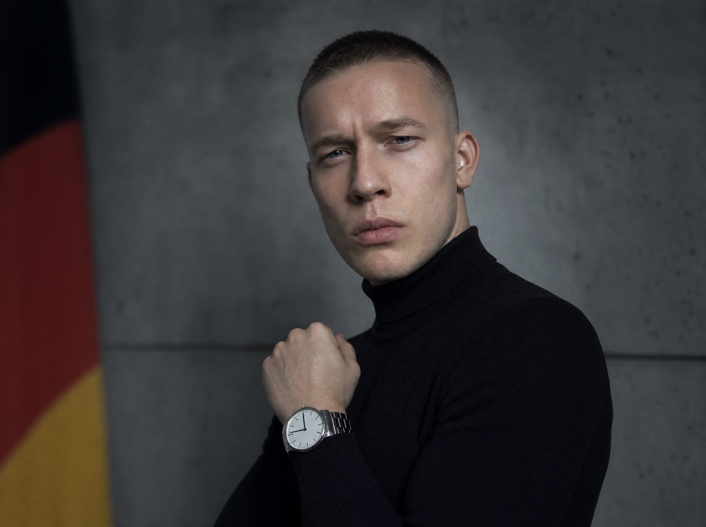 Leo in a black turtleneck against a concrete wall, German flag in negative space, watch on wrist